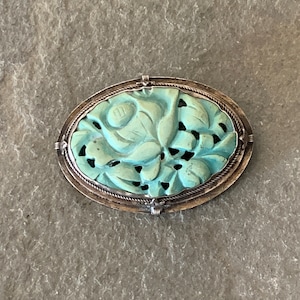 May include: A silver and turquoise brooch with a carved floral design. The brooch is oval shaped and has a detailed floral pattern.
