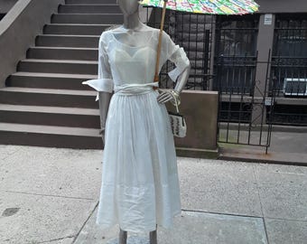 May include: A white dress with a fitted bodice and a full skirt. The dress is worn by a mannequin with a floral patterned parasol and a turban-style head covering.