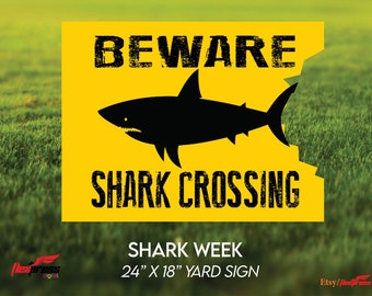 May include: A yellow yard sign with a black silhouette of a shark and the text "BEWARE SHARK CROSSING". The sign also includes the text "SHARK WEEK 24" x 18" YARD SIGN".