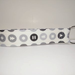 May include: A white fabric keychain with a repeating pattern of gray and black circles containing music symbols. The keychain has a silver metal ring.
