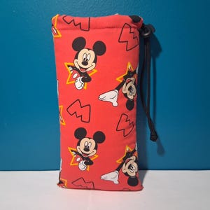 May include: Red glasses case featuring a repeating pattern of Mickey Mouse, yellow stars, and abstract shapes. The case has a black drawstring closure. The design is a playful and colorful accessory for eyewear.