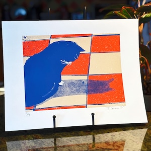 May include: A blue cat print on white paper. The cat is silhouetted against a red, white, and blue checkered background. The print is displayed on a black stand. The print is signed and numbered 3/14.