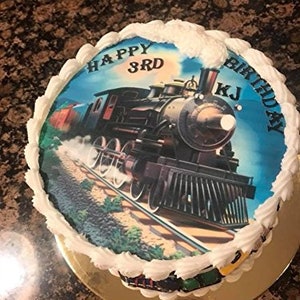 May include: A round birthday cake with white frosting and a train image. The image features a vintage steam engine on tracks, with the words "Happy 3rd Birthday KJ" written on the cake. The cake sits on a gold-colored cake board.