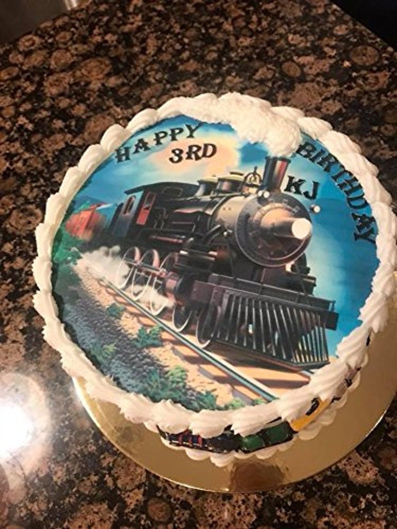 May include: A round birthday cake with white frosting and a train image. The image features a vintage steam engine on tracks, with the words "Happy 3rd Birthday KJ" written on the cake. The cake sits on a gold-colored cake board.