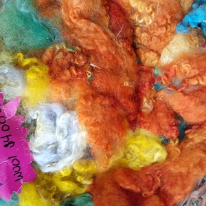 May include: A basket filled with colorful wool roving in shades of orange, yellow, green, blue, and white. The sign reads "Wool $4.00/100g" and "Wensleydale".
