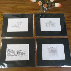 May include: Four black framed prints of ink drawings of buildings. The drawings are in black and white and are all different styles. The prints are all the same size and are hanging on a wooden wall.