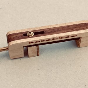 May include: A wooden clamp with a brass screw and accents. The clamp is made of light-colored wood with a dark wood inlay. The text "Hewn from the Mountain" is engraved on the side. The clamp is designed for woodworking or crafts.