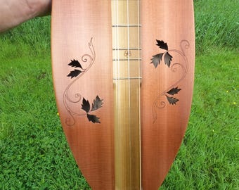 Op de afbeelding: Een houten dulcimer met een lichtbruine afwerking. Het instrument heeft een toets en snaren, met decoratieve blad- en wijnstokuitsparingen op de body. De dulcimer wordt tegen een achtergrond van groen gras gehouden.