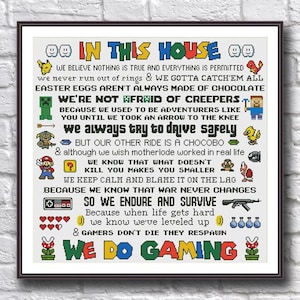 May include: Framed cross-stitch artwork with the text "IN THIS HOUSE" and various gaming-related phrases and pixelated graphics. The artwork is in a black frame and is displayed against a white brick wall.
