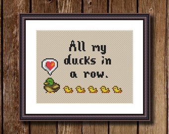 May include: Cross-stitch pattern featuring a green duck with a red heart speech bubble above it, and the text "All my ducks in a row."  Seven yellow ducklings are lined up below the text.