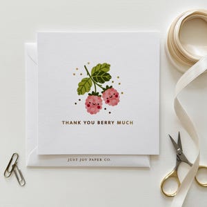 May include: A white greeting card with the text "THANK YOU BERRY MUCH" and an illustration of two pink raspberries with smiling faces. The card is on a white envelope, with a gold ribbon, scissors, and paper clips.