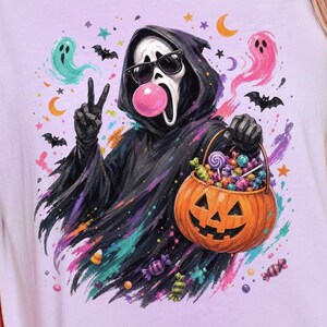 May include: A light purple t-shirt featuring a graphic of a hooded figure in a black robe wearing sunglasses and blowing a pink bubble. The figure holds a jack-o'-lantern bucket filled with sweets, surrounded by colourful ghosts, bats, and stars.