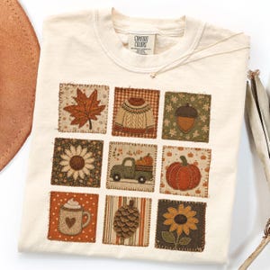 May include: A cream-colored t-shirt featuring a grid of nine square patches with autumn-themed designs, including a maple leaf, a knit sweater, an acorn, sunflowers, a truck carrying pumpkins, a large pumpkin, a coffee mug, and a pinecone.