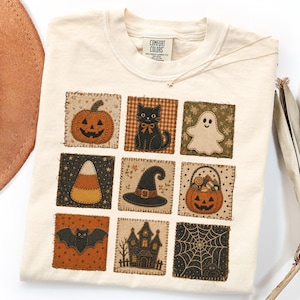 May include: A cream-colored t-shirt featuring a grid of nine square patches with Halloween-themed designs, including a pumpkin, black cat, ghost, candy corn, witch hat, jack-o'-lantern basket, bat, haunted house, and spider web.