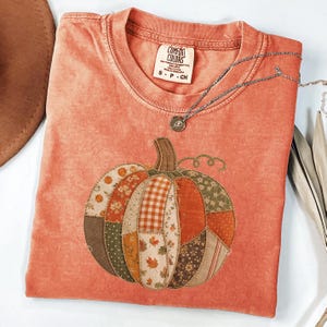 May include: A coral-coloured t-shirt featuring a large, central pumpkin graphic composed of various fabric-like patches in shades of orange, green, and brown with floral, plaid, and leaf patterns. The shirt has a Comfort Colors brand label.