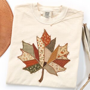 May include: A cream-colored crew neck t-shirt featuring a large, central maple leaf graphic composed of various fabric patches in shades of brown, orange, green, and cream. The patches are joined by visible stitching, and the shirt has a Comfort Colors label.