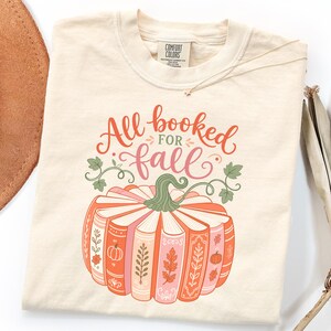 Könnte beinhalten: Ein cremefarbenes T-Shirt mit einer Grafik eines Kürbisses, der aus gestapelten Büchern besteht. Der Text All booked for fall ist in orangefarbener und rosafarbener Schreibschrift gedruckt. Das Design enthält grüne Ranken sowie kleine Blätter.