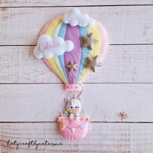 Può includere: Un pallone aerostatico rosa e giallo con strisce arcobaleno e una nuvola bianca con il nome Celeste scritto su di essa. Il pallone ha tre stelle dorate scintillanti attaccate ad esso. Un unicorno è seduto in un cestino rosa attaccato alla parte inferiore del pallone.