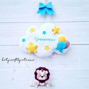 Può includere: Una nuvola di feltro bianco con stelle gialle e palloncini blu, con il nome "Gianmarco" scritto su di essa, è appesa a un fiocco blu. Un leone marrone con una corona siede su un'altalena di legno.