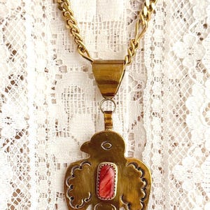 May include: A gold-toned metal necklace featuring a bird-shaped pendant with a central rectangular red stone inlay. The pendant displays etched decorative patterns along its wings and tail, suspended from a gold-toned chain with flat links.