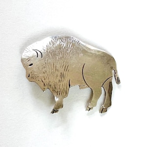 Puede incluir: Un broche de metal plateado con forma de bisonte. El broche tiene la forma de un bisonte, con una textura detallada en el cuerpo y un acabado liso. El bisonte se representa de perfil, con cuernos y patas visibles. Probablemente sea una joya.