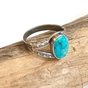 May include: A silver ring featuring an oval turquoise stone. The ring has a decorative design on the band with etched patterns. The turquoise stone is a vibrant blue-green color, set in a silver bezel. The ring is displayed on a piece of wood.