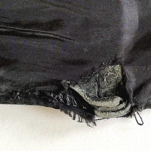 Op de afbeelding: Close-up van een gescheurde zwarte stof, met een beschadigd gebied met gerafelde randen en een gedeeltelijk blootgestelde, getextureerde laag eronder. De stof lijkt een duurzaam materiaal te zijn, mogelijk voor bovenkleding.