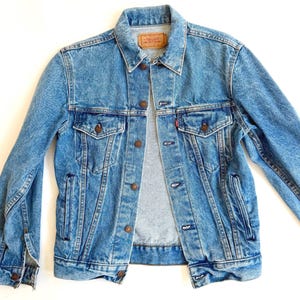 May include: Light blue denim jacket with button closures, two chest pockets, and a small red tag. The jacket has a classic design with a collared neckline and buttoned cuffs. The Levi's brand label is visible.