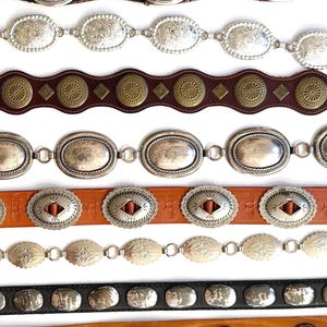Op de afbeelding: Een collectie van acht riemen die horizontaal zijn gerangschikt. De riemen hebben diverse ontwerpen, waaronder zilverkleurige ovale concho's, messingkleurige ronde studs, leren banden in zwart, bruin en beige, en gedetailleerde metalen afwerkingen.