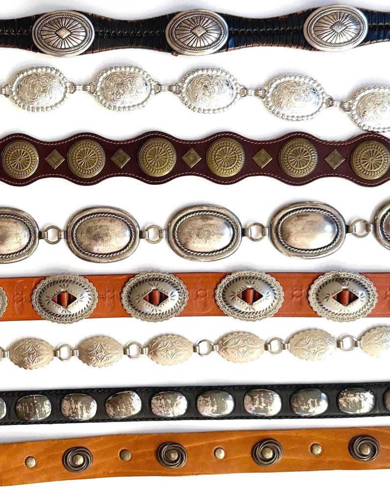 Op de afbeelding: Een collectie van acht riemen die horizontaal zijn gerangschikt. De riemen hebben diverse ontwerpen, waaronder zilverkleurige ovale concho's, messingkleurige ronde studs, leren banden in zwart, bruin en beige, en gedetailleerde metalen afwerkingen.