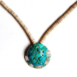 May include: A turquoise and silver pendant necklace. The pendant is a round shape with a mosaic of turquoise stones set in a silver frame. The necklace chain is made of small, off-white shell beads.