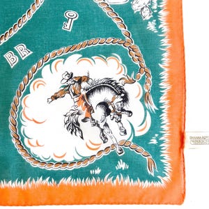 K&ouml;nnte beinhalten: Quadratisches, t&uuml;rkisfarbenes Halstuch mit orangefarbenem Rand. Das Design zeigt einen Cowboy, der auf einem wilden Pferd in einer wei&szlig;en Wolke reitet, umgeben von einem Seilmuster. Die Buchstaben "BR" sind in der oberen linken Ecke zu sehen.