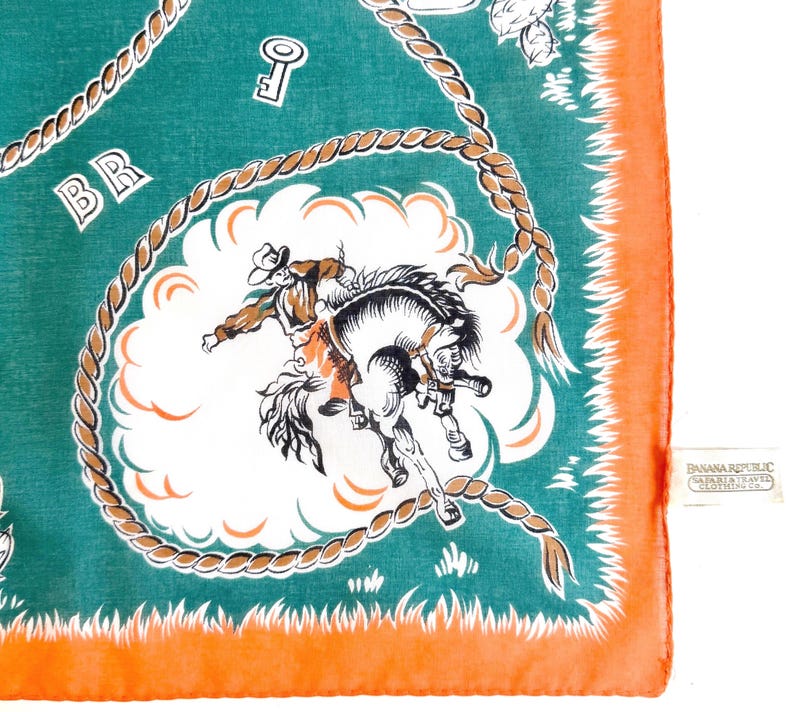 K&ouml;nnte beinhalten: Quadratisches, t&uuml;rkisfarbenes Halstuch mit orangefarbenem Rand. Das Design zeigt einen Cowboy, der auf einem wilden Pferd in einer wei&szlig;en Wolke reitet, umgeben von einem Seilmuster. Die Buchstaben "BR" sind in der oberen linken Ecke zu sehen.