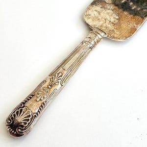 May include: Close-up of a vintage silver cake server. The handle features ornate floral and scrollwork designs. The blade is rectangular with a slightly rounded edge. The server has a tarnished, antique appearance.