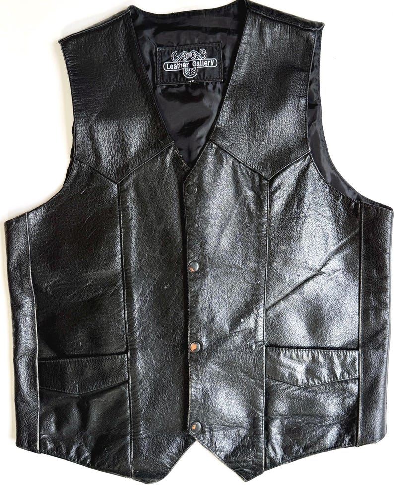 Op de afbeelding: Zwarte leren vest met een V-hals, twee voorzakken en drie knoopsluitingen. Het vest heeft een satijnen voering en een label met de tekst "Leather Gallery". Een klassieke stijl.