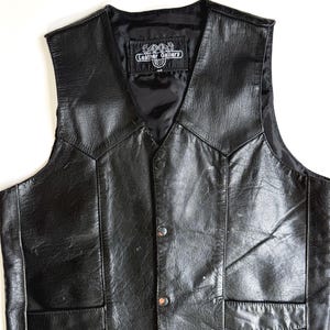 Op de afbeelding: Zwarte leren vest met een V-hals en twee voorzakken. Het vest heeft een knoopsluiting en een satijnen voering. Het label luidt "Leather Gallery".