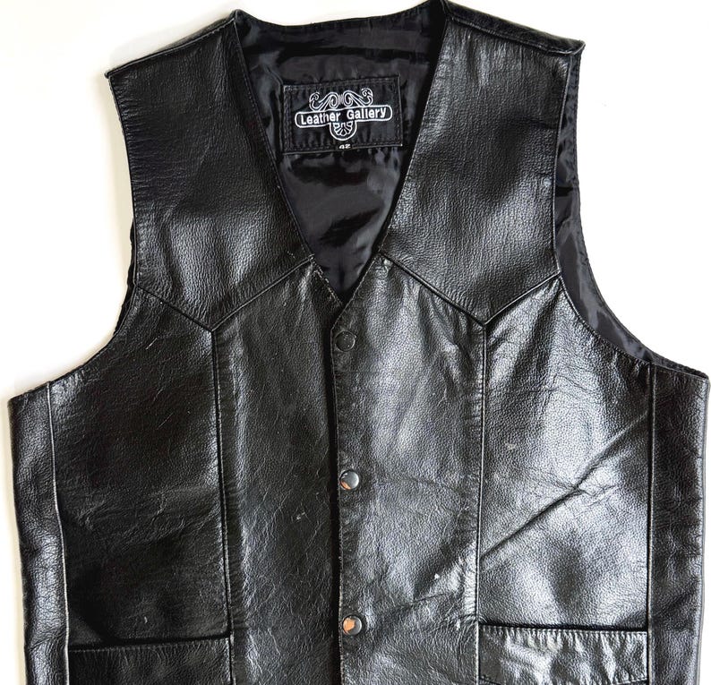 Op de afbeelding: Zwarte leren vest met een V-hals en twee voorzakken. Het vest heeft een knoopsluiting en een satijnen voering. Het label luidt "Leather Gallery".