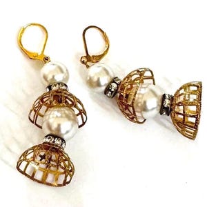 May include: A pair of gold-toned dangle earrings featuring white faux pearls, small clear rhinestone spacers, and intricate gold-toned metal cage-like bell shapes. The earrings are attached to gold-toned lever-back closures for secure wear.