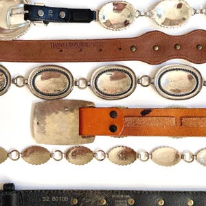Op de afbeelding: Een collectie van zes horizontaal gerangschikte riemen. Het assortiment omvat donkerbruin leer, zwart leer met zilverkleurige ovale concho-accenten, een beige leren Banana Republic-riem, een zilverkleurige kettingriem, een beige leren riem met een grote rechthoekige gesp en een zwarte leren riem met een gele su&egrave;de onderkant.