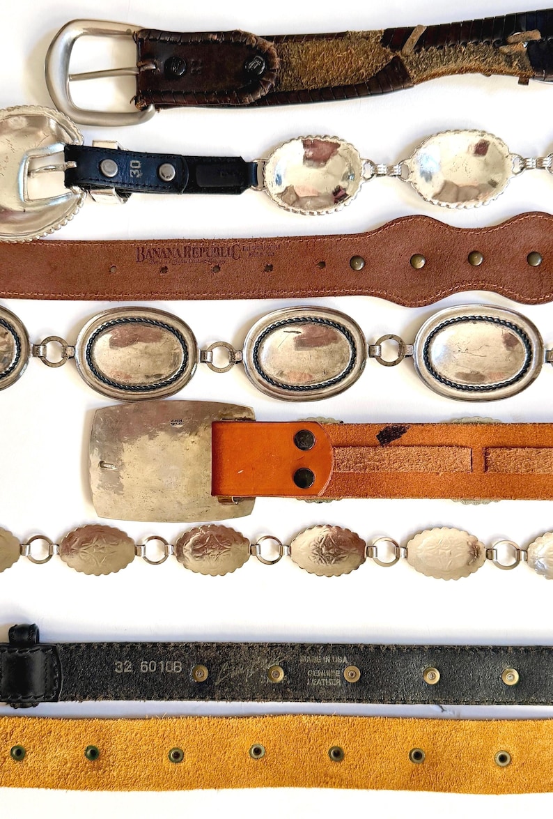 Op de afbeelding: Een collectie van zes horizontaal gerangschikte riemen. Het assortiment omvat donkerbruin leer, zwart leer met zilverkleurige ovale concho-accenten, een beige leren Banana Republic-riem, een zilverkleurige kettingriem, een beige leren riem met een grote rechthoekige gesp en een zwarte leren riem met een gele su&egrave;de onderkant.