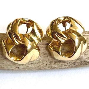 May include: A pair of gold-coloured, chunky chain link earrings. The earrings are made of a shiny, polished metal and have a post backing. The earrings are displayed on a piece of driftwood against a white background.