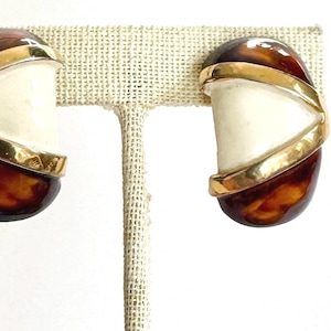 May include: A pair of vintage clip-on earrings. The earrings have a cream-coloured centre, framed by a gold-tone metal band. The bottom half is a tortoiseshell pattern. The earrings are displayed on a neutral-coloured earring stand.