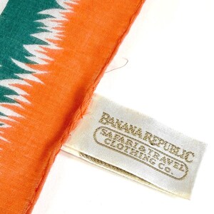 K&ouml;nnte beinhalten: Nahaufnahme eines orangefarbenen und teal-farbenen Schals mit einem wei&szlig;en und teal-farbenen Muster. Der Schal hat ein Etikett mit der Aufschrift "Banana Republic Safari & Travel Clothing Co."