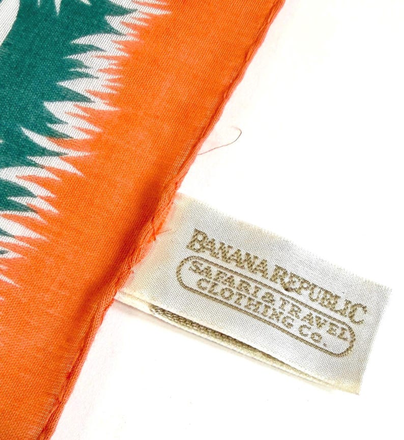 K&ouml;nnte beinhalten: Nahaufnahme eines orangefarbenen und teal-farbenen Schals mit einem wei&szlig;en und teal-farbenen Muster. Der Schal hat ein Etikett mit der Aufschrift "Banana Republic Safari & Travel Clothing Co."