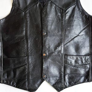 Op de afbeelding: Een zwart leren vest met een klassiek ontwerp. Het vest heeft een V-hals, twee voorzakken en een knoopsluiting. Het leer heeft een licht versleten uiterlijk.