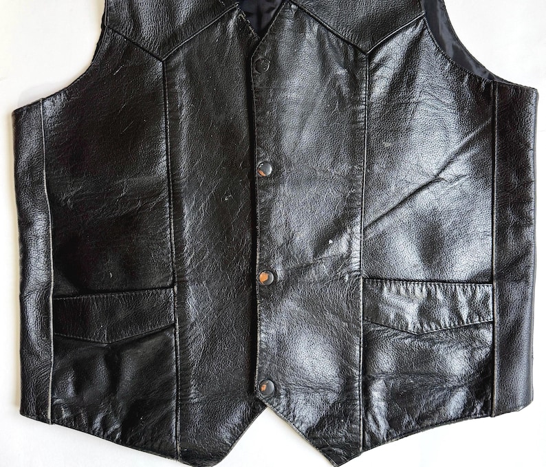 Op de afbeelding: Een zwart leren vest met een klassiek ontwerp. Het vest heeft een V-hals, twee voorzakken en een knoopsluiting. Het leer heeft een licht versleten uiterlijk.
