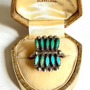 Puede incluir: Anillo de plata vintage con piedras turquesas y verdes, presentado en un joyero dorado. El anillo presenta un diseño rectangular con múltiples piedras ovaladas en colores alternos. El joyero está forrado con tela color crema.