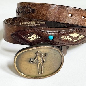 以下が含まれることがあります： カウボーイの姿が描かれた真鍮製のバックルが付いた、茶色の革ベルト。 ベルトには装飾的なステッチ、パイソンとターコイズのアクセント、そして「SOUTHWICK LEATHERS PYTHON/TURQUOISE」の文字が施されています。