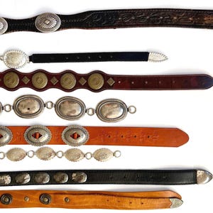 Op de afbeelding: Een collectie van acht riemen horizontaal gerangschikt, met diverse ontwerpen waaronder donkerbruin leer met zilveren concho's, zwart leer met zilveren accenten, lichtbruin leer met messingkleurige ronde studs en zilverkleurige metalen schakelkettingen.