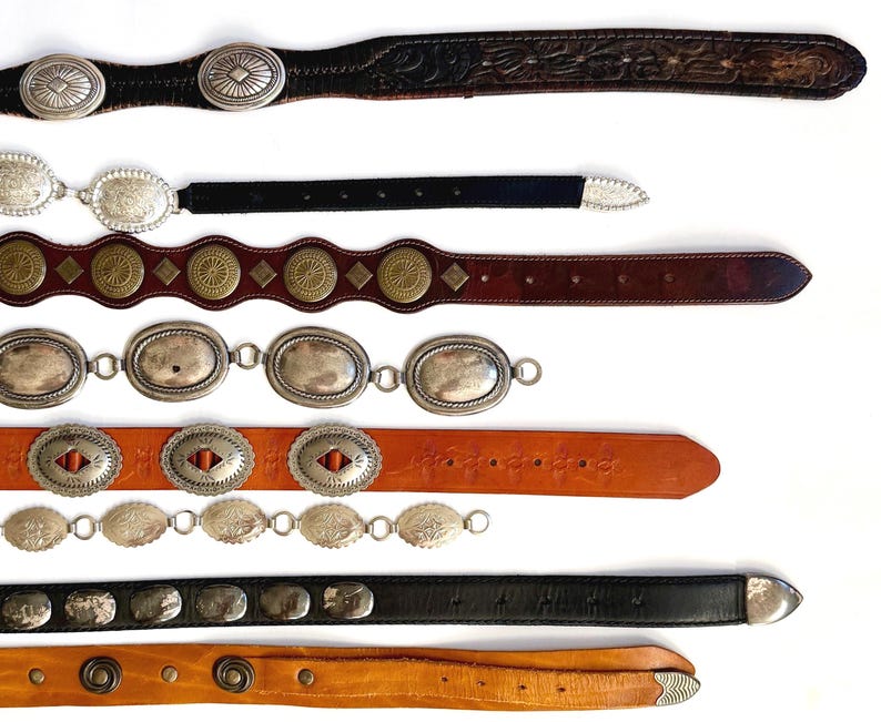 Op de afbeelding: Een collectie van acht riemen horizontaal gerangschikt, met diverse ontwerpen waaronder donkerbruin leer met zilveren concho's, zwart leer met zilveren accenten, lichtbruin leer met messingkleurige ronde studs en zilverkleurige metalen schakelkettingen.